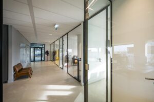 Moderne kantoorhal op Rietlandpark met glazen kantoordeuren, een leren bankje en zicht op meerdere werkplekken.