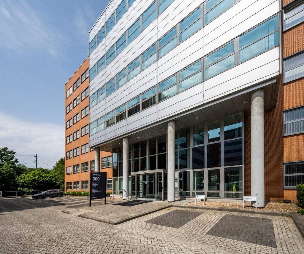 Hoofdingang van kantoorgebouw Entrada 300 met grote glazen gevel, pilaren en een informatiebord bij de oprit.