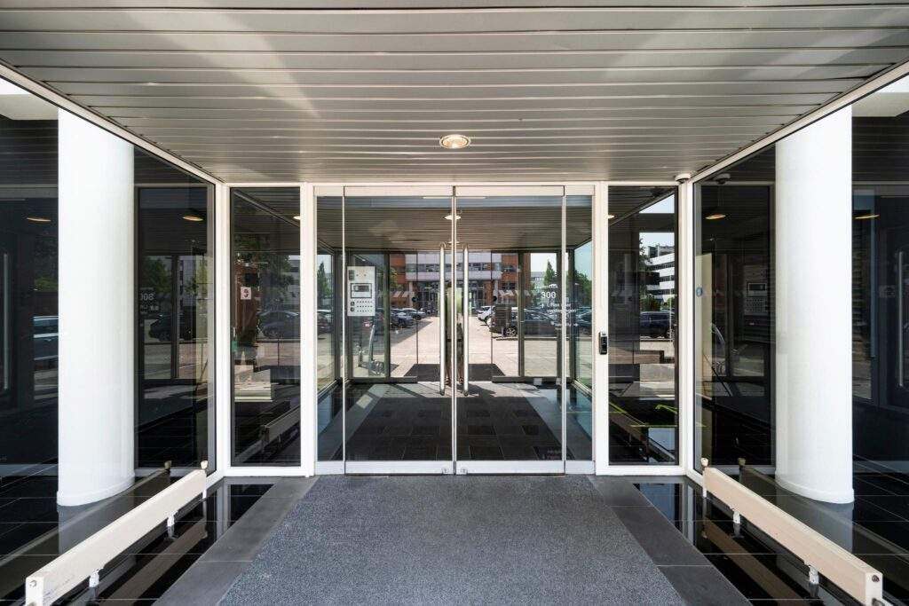 Glazen entree van een modern kantoorgebouw met dubbele deuren en zicht op de parkeerplaats buiten.