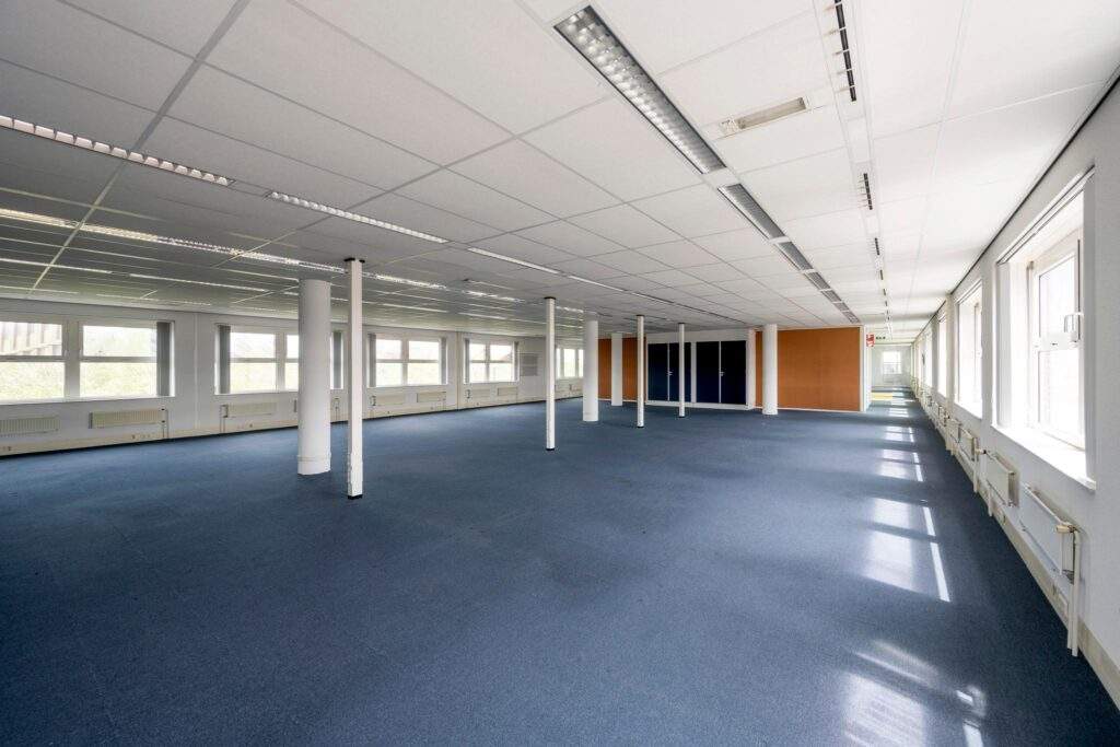 Ruime, lege kantoorruimte met blauw tapijt, systeemplafond en grote ramen langs de rechterzijde.