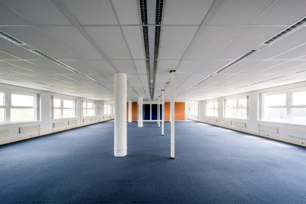 Lege, lichte kantoorruimte met blauwe vloerbedekking, witte pilaren en grote ramen aan beide zijden.