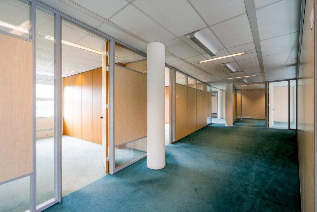 Gang met gesloten en deels geopende kantoorkamers met houten scheidingswanden en blauw tapijt.