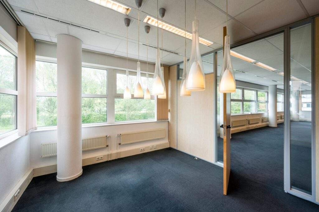 Modern, licht kantoorpand met grote ramen, glazen wanden en hanglampen.