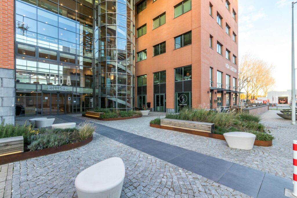 Entree van het VIDA Gebouw aan de Kabelweg met moderne glazen gevel, beplanting en zitgelegenheid.