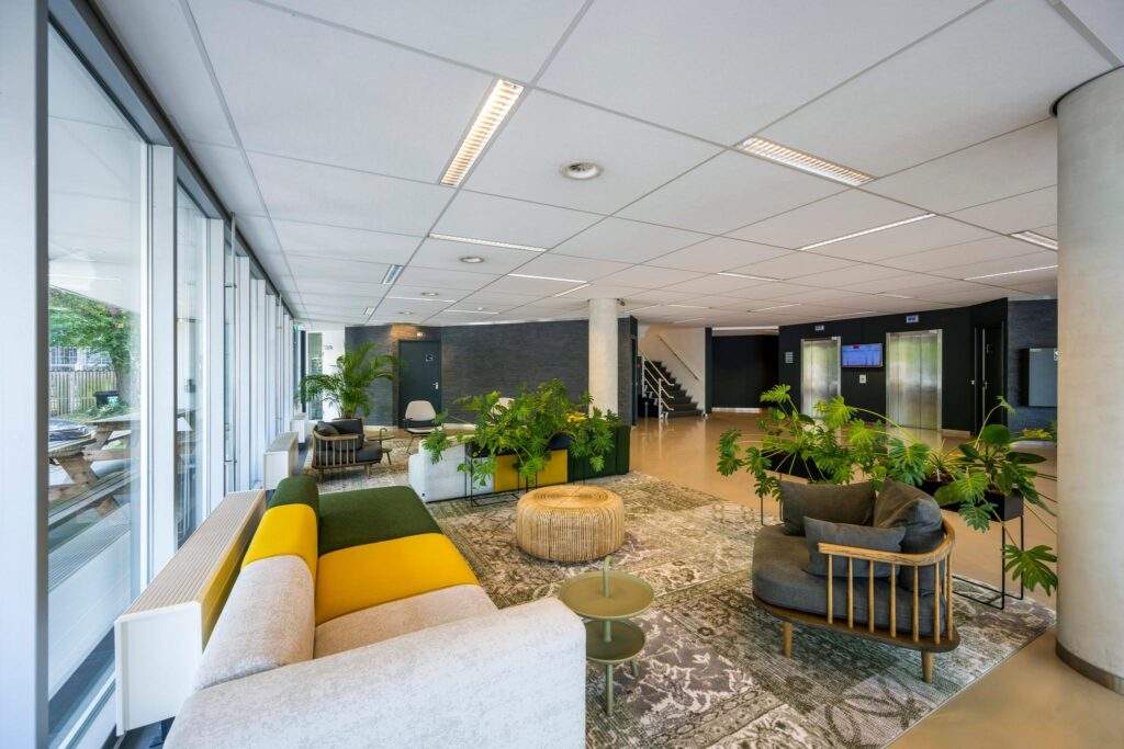 Moderne en kleurrijke ontvangstruimte in het kantoor aan de Hogehilweg, met comfortabele zitplaatsen, veel groen en liften op de achtergrond.
