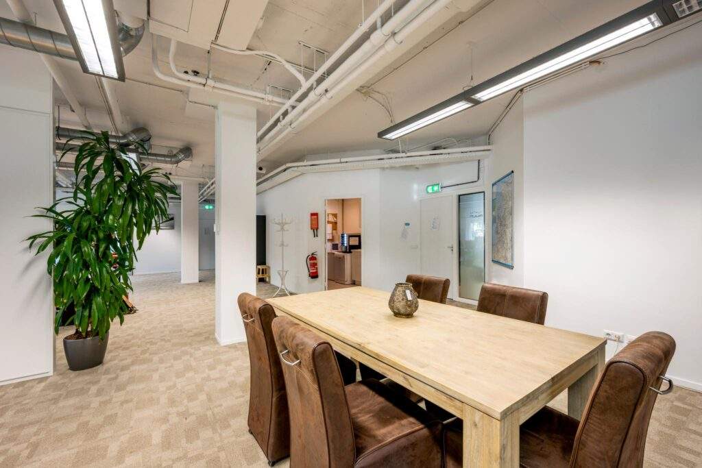 Moderne kantoorruimte aan de Hogehilweg met een houten vergadertafel, zes leren stoelen en zicht op een pantry en kantoorruimte.