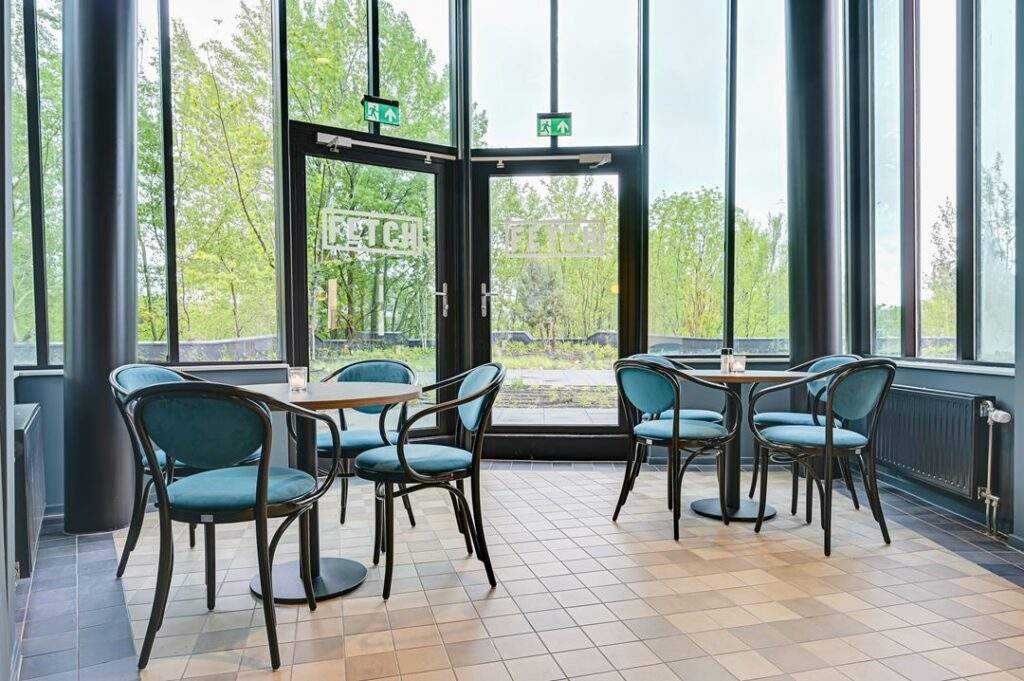 Modern ingerichte zitruimte met blauwe stoelen en ronde tafels bij de ingang van gebouw Kingsfordweg, met uitzicht op groen buitengebied.