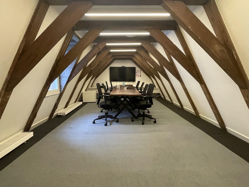 Modern conference room in an attic with exposed wooden beams, a long table, office chairs, and a large screen.