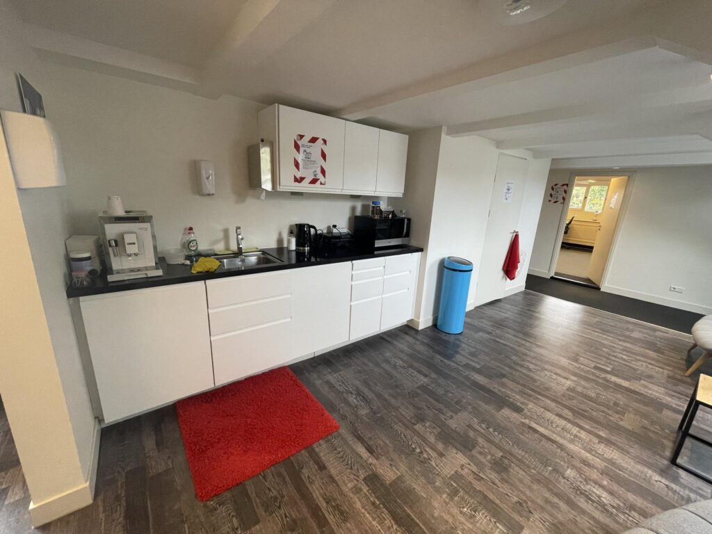 Modern shared kitchenette with white cabinets, coffee machine, microwave, and a red carpet on a dark wooden floor in a communal workspace at Herengracht.