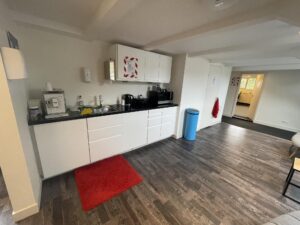 Modern shared kitchenette with white cabinets, coffee machine, microwave, and a red carpet on a dark wooden floor in a communal workspace at Herengracht.