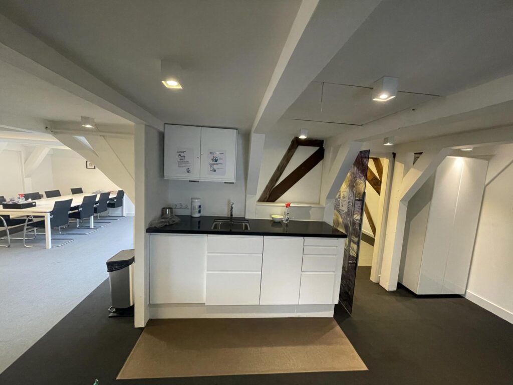 Modern office kitchenette with a sink and white cabinets, adjacent to a conference room with a long table and chairs.