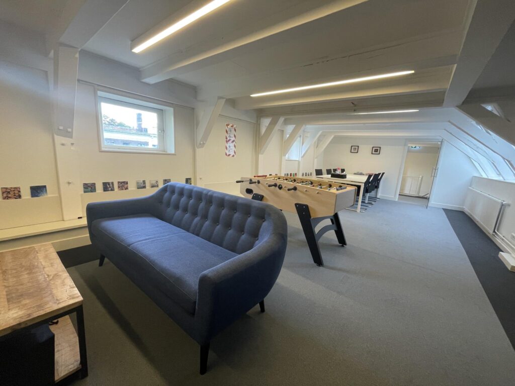 Modern office lounge at Herengracht 174 featuring a blue sofa, foosball table, and meeting area under a slanted ceiling.