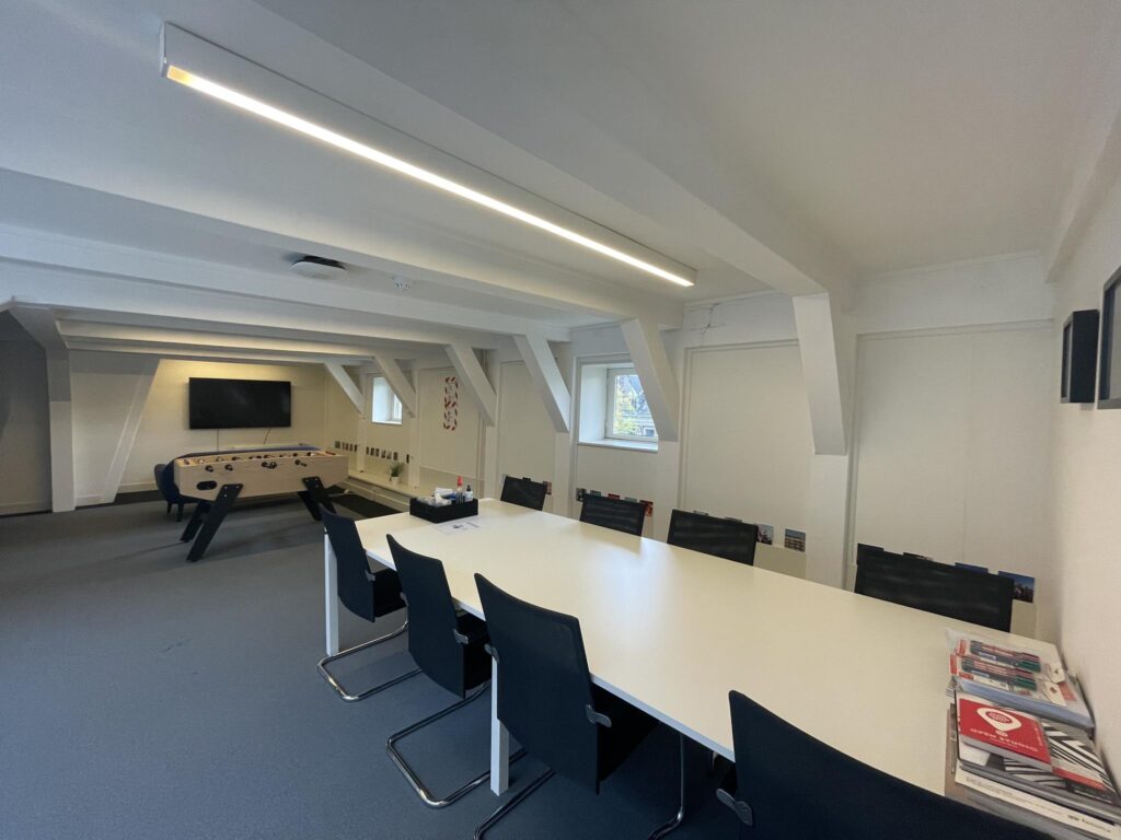 Modern meeting room with a long white conference table, black chairs, a foosball table, and a wall-mounted TV.