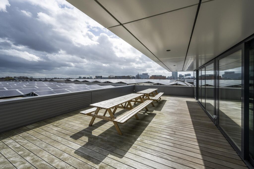 Dakterras met houten picknicktafels en uitzicht op zonnepanelen en de skyline van Amsterdam-Dynamostraat.