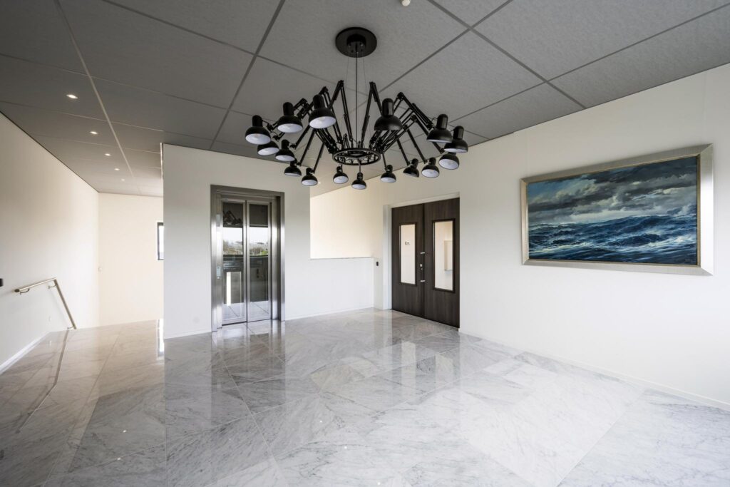 Moderne hal met marmeren vloer, opvallende zwarte plafondlamp, een lift, dubbele donkere deuren en een schilderij van een woeste zee aan de muur.