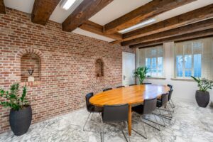 Vergaderruimte aan de Herengracht met een ovale houten tafel, bakstenen muur, marmeren vloer en zichtbare houten plafondbalken.