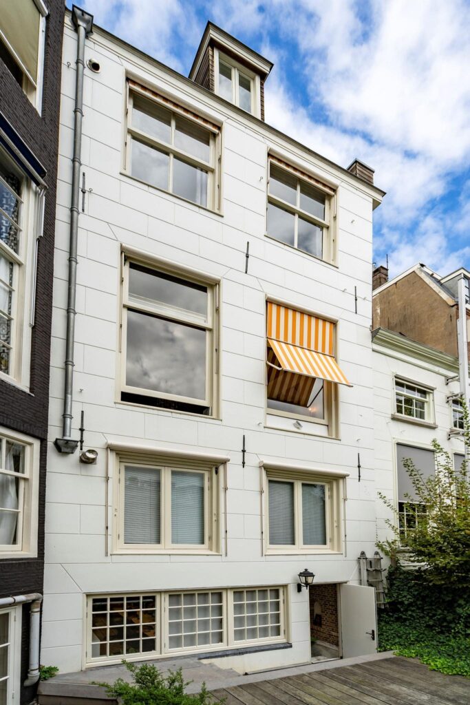 Witgepleisterde gevel van een grachtenpand aan de Herengracht met grote ramen en een geel-wit gestreept zonnescherm.