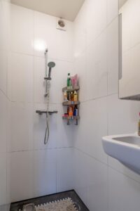 Douche met witte wandtegels, een handdouche, hoekrek met verzorgingsproducten en een wastafel in een badkamer aan de Herengracht.