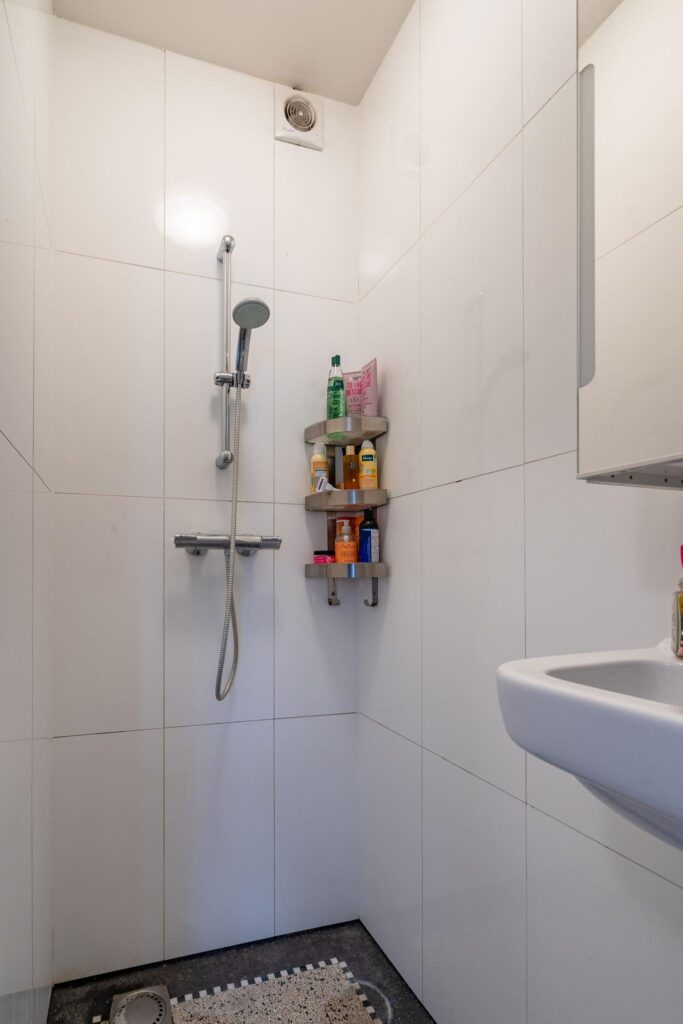 Douche met witte wandtegels, een handdouche, hoekrek met verzorgingsproducten en een wastafel in een badkamer aan de Herengracht.