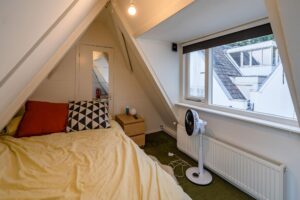 Zolderkamer aan de Herengracht met een schuin dak, een bed, ventilator en uitzicht op omliggende daken.