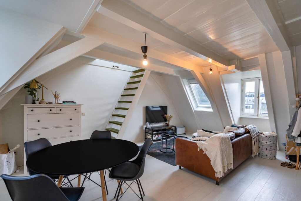 Zolderappartement aan de Herengracht met schuine houten balken, een leren bank, eettafel en open trap naar een hoger gelegen verdieping.