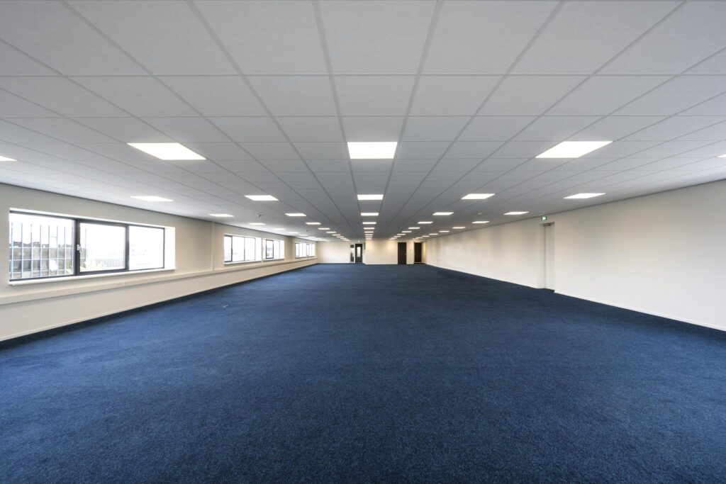 Spacious empty office space with blue carpet, white walls, and large windows on Dynamostraat.