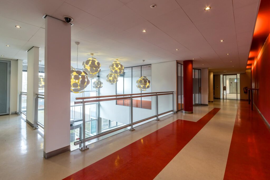 Moderne binnenruimte van een gang in een kantoor- of zorggebouw aan de Lemelerbergweg, met opvallende rode vloer, glazen balustrade en bolvormige hanglampen.