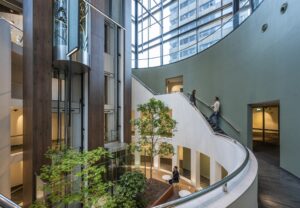 Moderne, lichte binnentuin met glazen liften, groene planten en een gebogen trap in Amstelplein-kantoorgebouw.