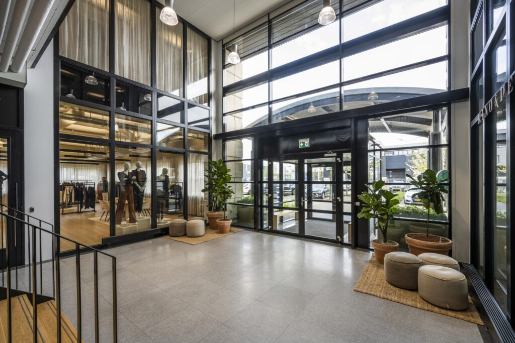 Modern office entrance with large glass windows, indoor plants, and a view into a stylish showroom.