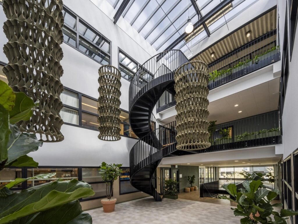 Modern atrium interior of a building on Singaporestraat with a black spiral staircase, large hanging sculptures, and abundant greenery.