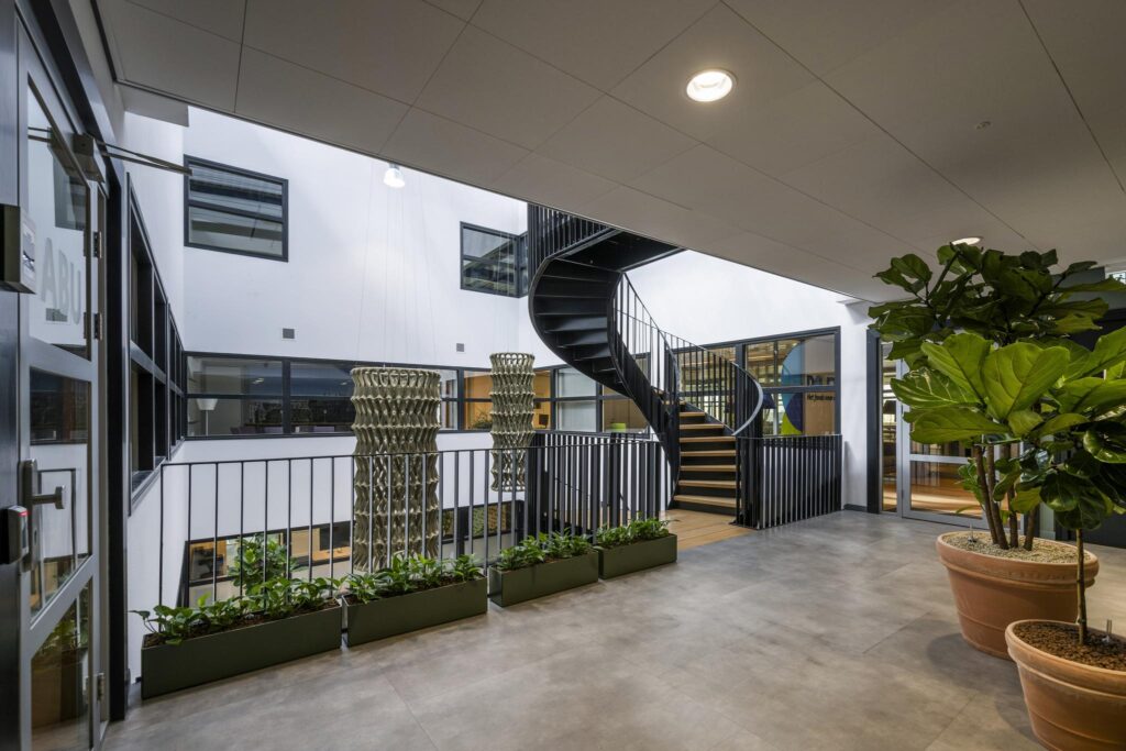 Modern indoor office courtyard with a black spiral staircase, large potted plants, and natural light.