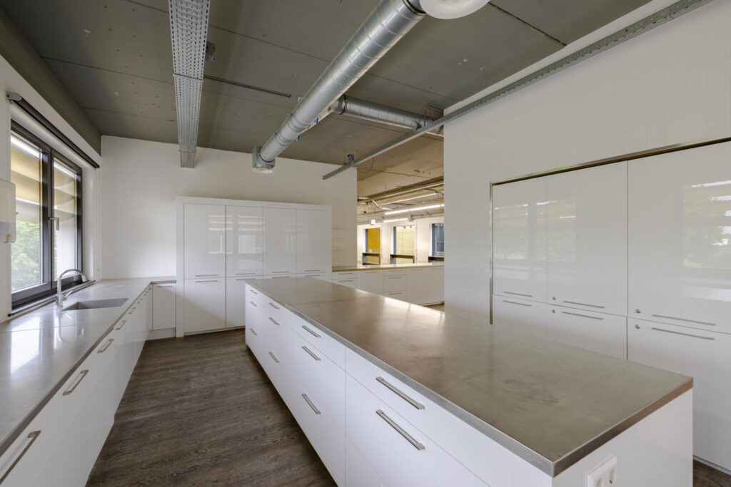Moderne, ruime keuken met glanzend witte kasten en roestvrijstalen werkbladen in een bedrijfs- of onderwijsomgeving aan de Laarderhoogtweg.