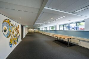 Moderne kantoorruimte met glazen wanden, wandkunst in felle kleuren en een houten tafel langs het raam.