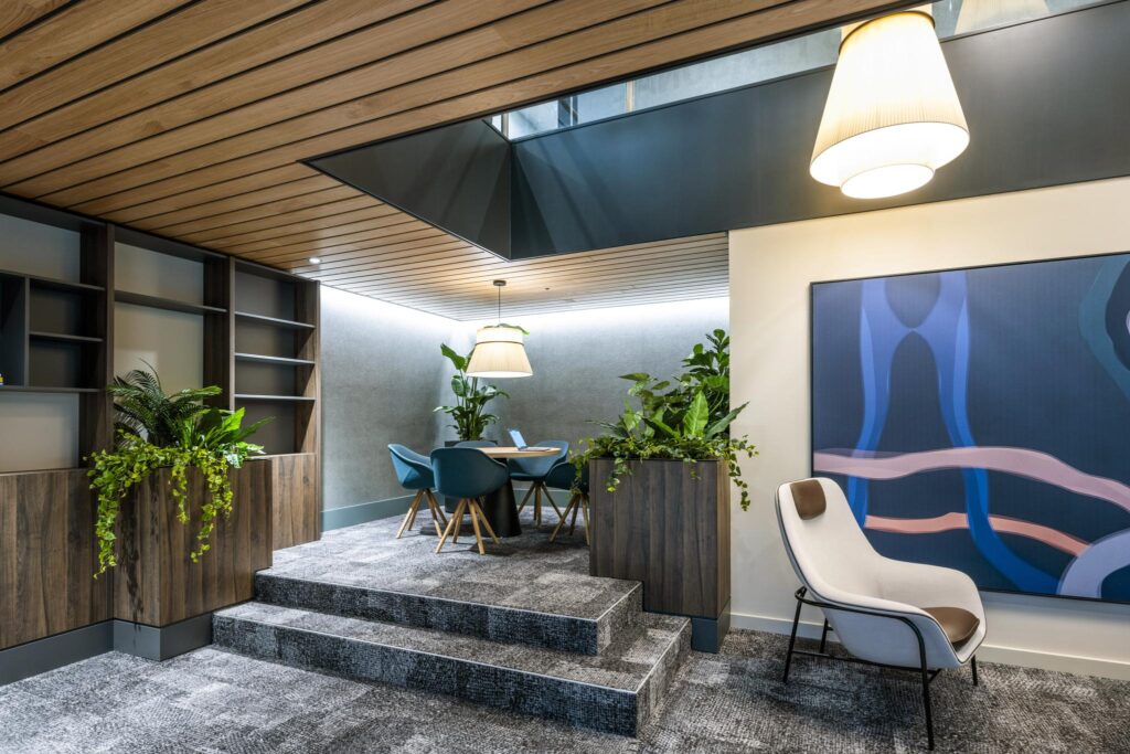 Modern office lounge with wooden ceiling, indoor plants, blue chairs, and contemporary artwork on the wall.