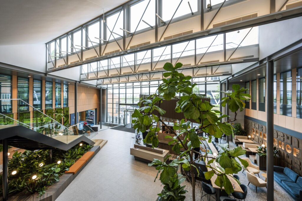 Modern lobby with large windows, indoor plants, seating areas, and a central staircase at Jachthavenweg.