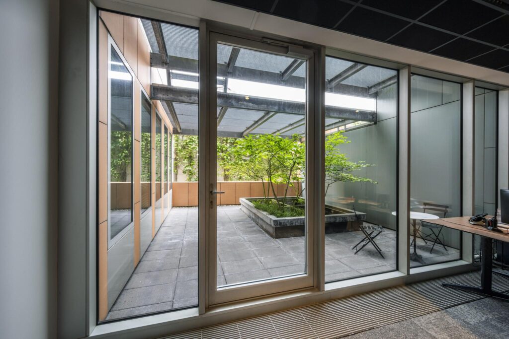 Glass door view of a modern courtyard with a green tree, patio tiles, and seating area at Jachthavenweg.
