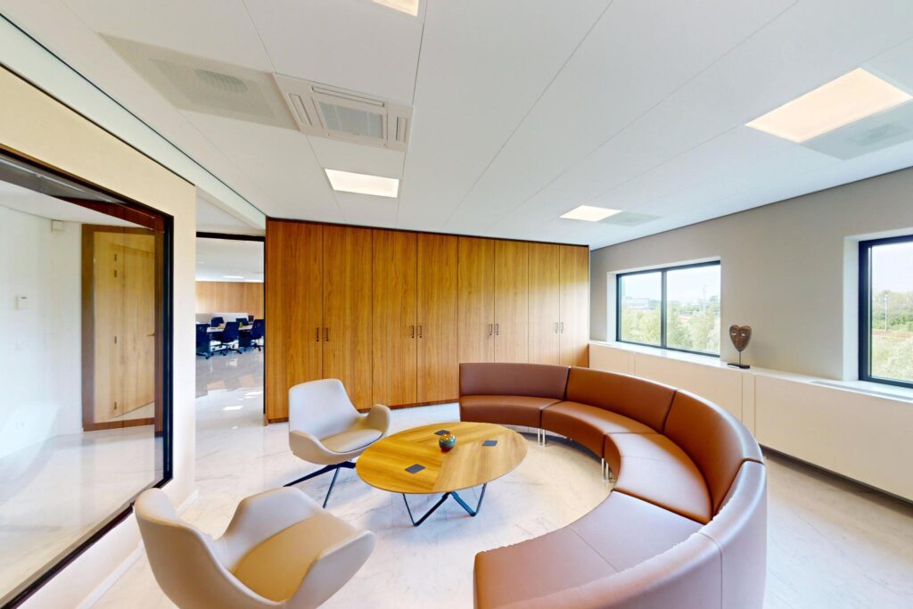 Modern office lounge with curved brown seating, two armchairs, a round wooden coffee table, and large windows.