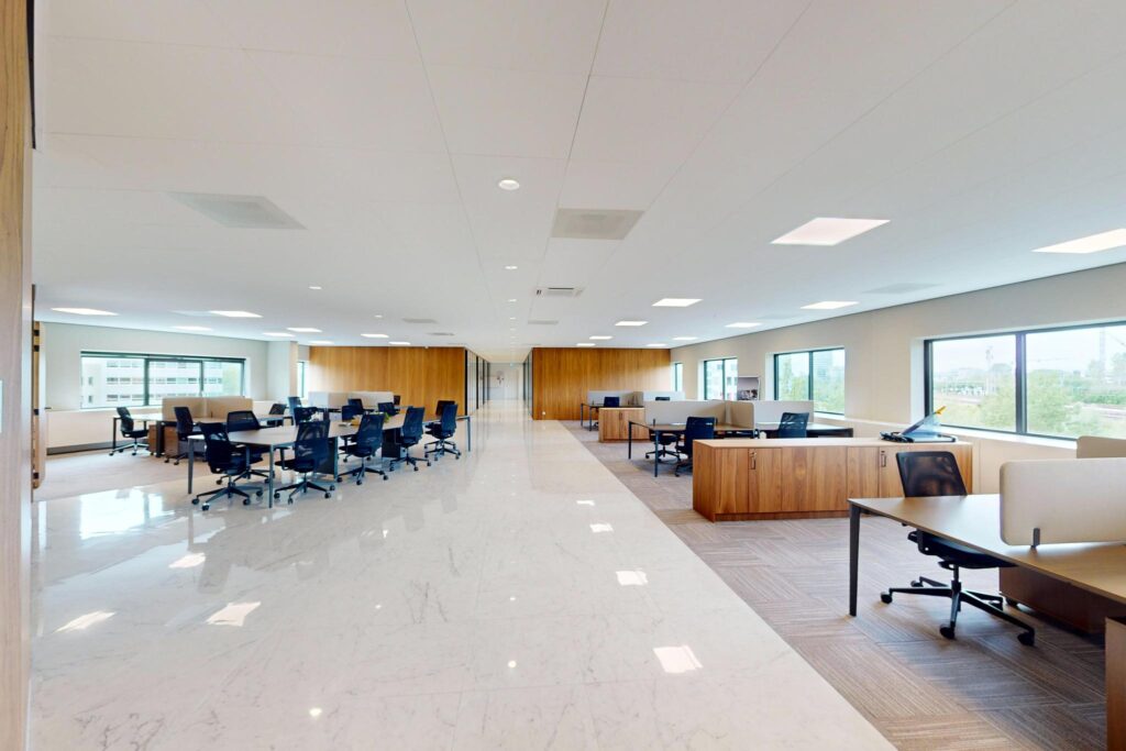 Modern open-plan office space with rows of desks, ergonomic chairs, and large windows providing natural light.