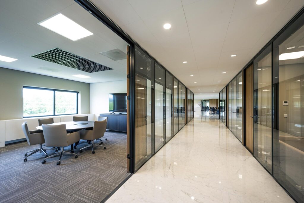 Modern office hallway with glass-walled meeting rooms and a conference table visible on the left.