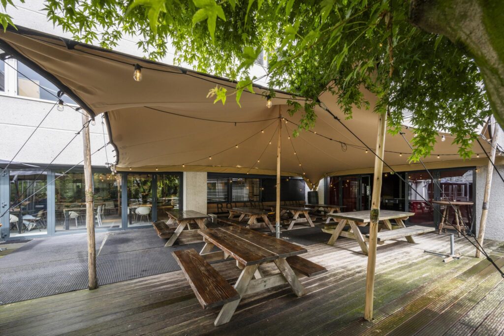 Outdoor seating area at Diemermeer with wooden picnic tables under a large beige canopy and string lights.