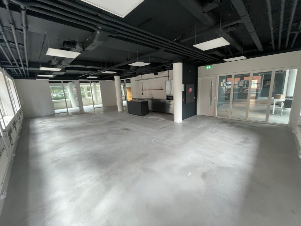 Spacious, modern industrial-style interior with concrete flooring, black ceiling, and a kitchenette area near large windows and glass doors.