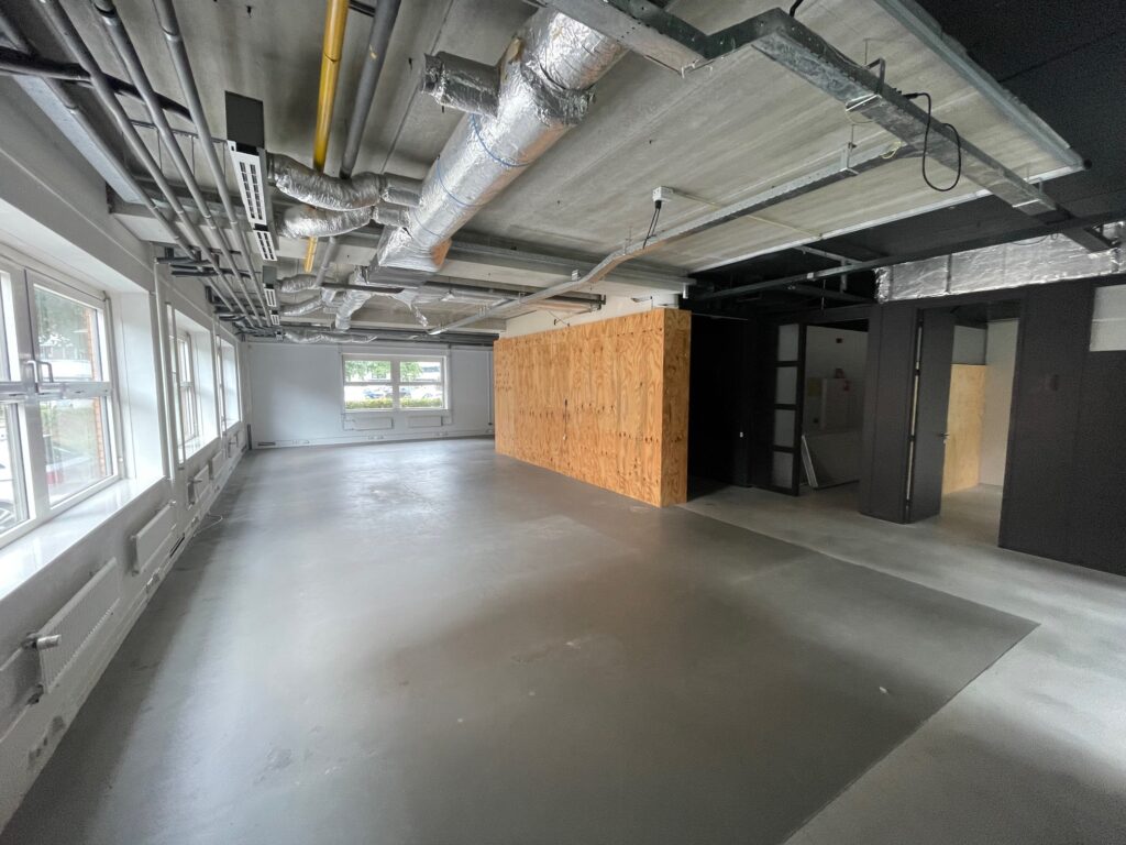 Industrial-style empty office space with exposed ductwork, large windows, and a wooden partition wall.