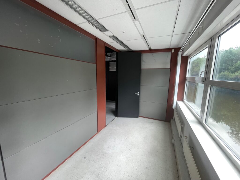 Small empty office space with grey panel walls, a black door, and large windows letting in natural light.