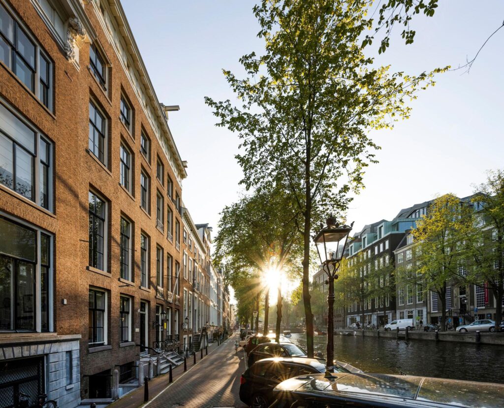 Zonsondergang aan de Herengracht met historische panden, bomen en geparkeerde auto's langs de gracht in Amsterdam.