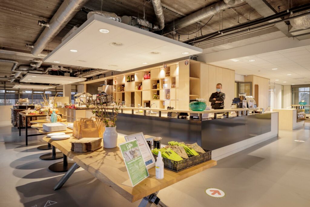 Modern ingerichte kantine aan de Karspeldreef met een open keuken, buffettafels en een medewerker achter de balie.