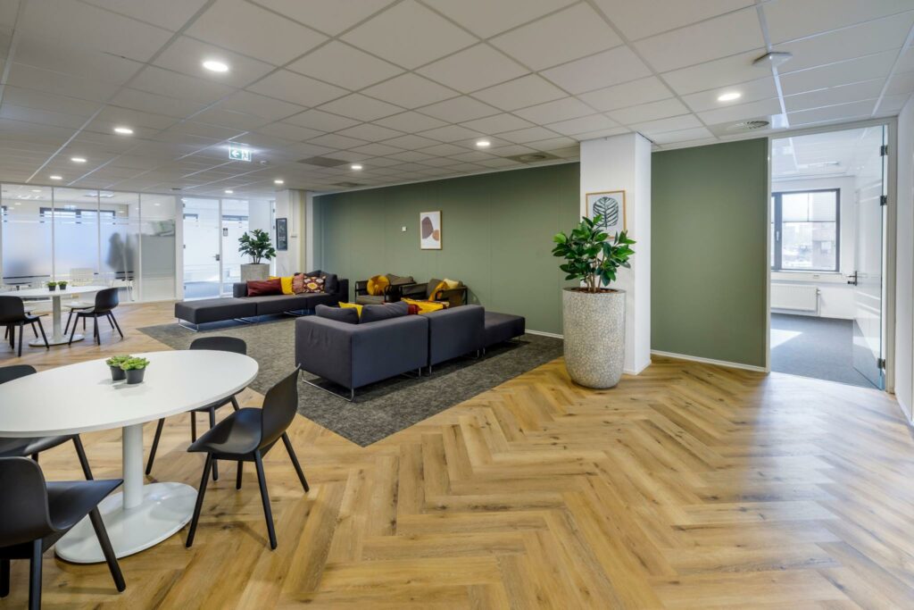 Moderne kantoorlounge aan de Karspeldreef met zithoeken, ronde tafels, houten visgraatvloer en groene accentmuur.