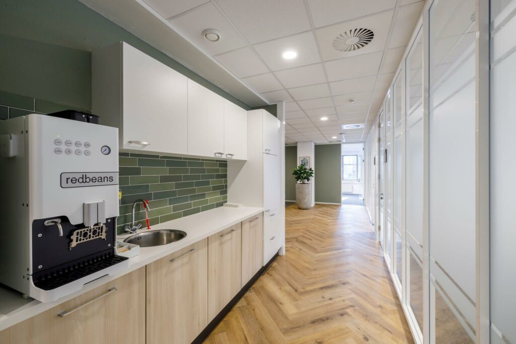 Moderne kantoorpantry aan de Karspeldreef met een koffiemachine, licht houten kastjes en groene wandtegels.