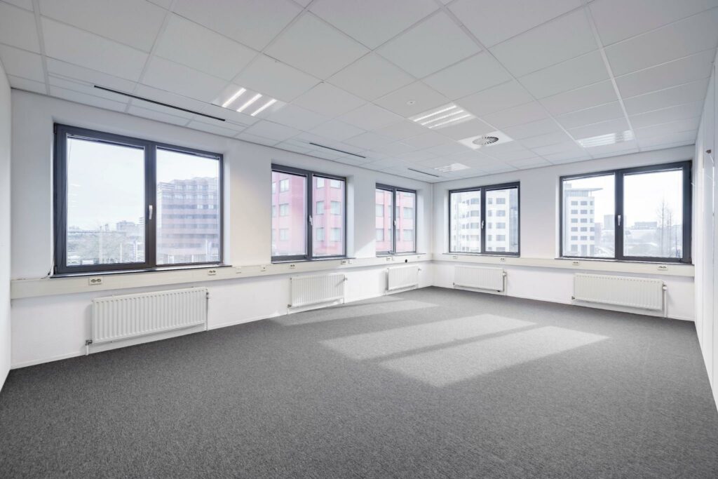 Lichte, lege kantoorruimte aan de Karspeldreef met grote ramen en uitzicht op omliggende kantoorgebouwen.