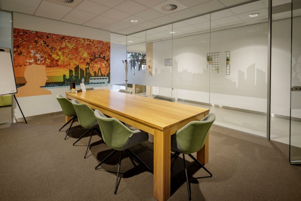 Vergaderruimte aan de Karspeldreef met een houten tafel, groene stoelen en een kleurrijke wanddecoratie met stadsgezicht en herfstbladeren.