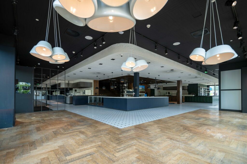 Modern interieur van een leeg restaurant op het Stadionplein met houten visgraatvloer, open keuken en grote hanglampen.